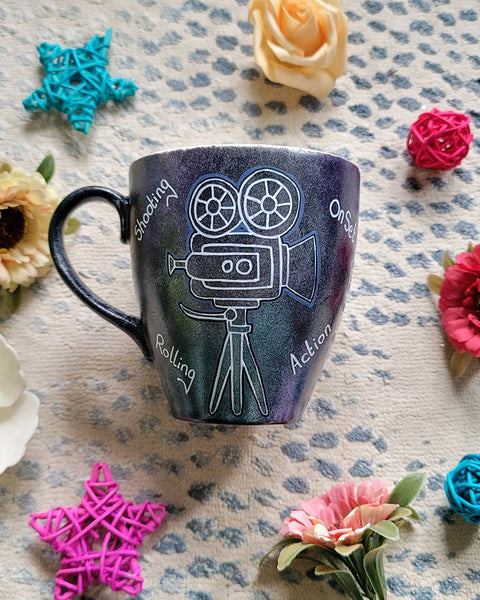 A Dark sky mug with a movie camera painted on it and words "Rolling. on set , Shooting, action" scattered all over