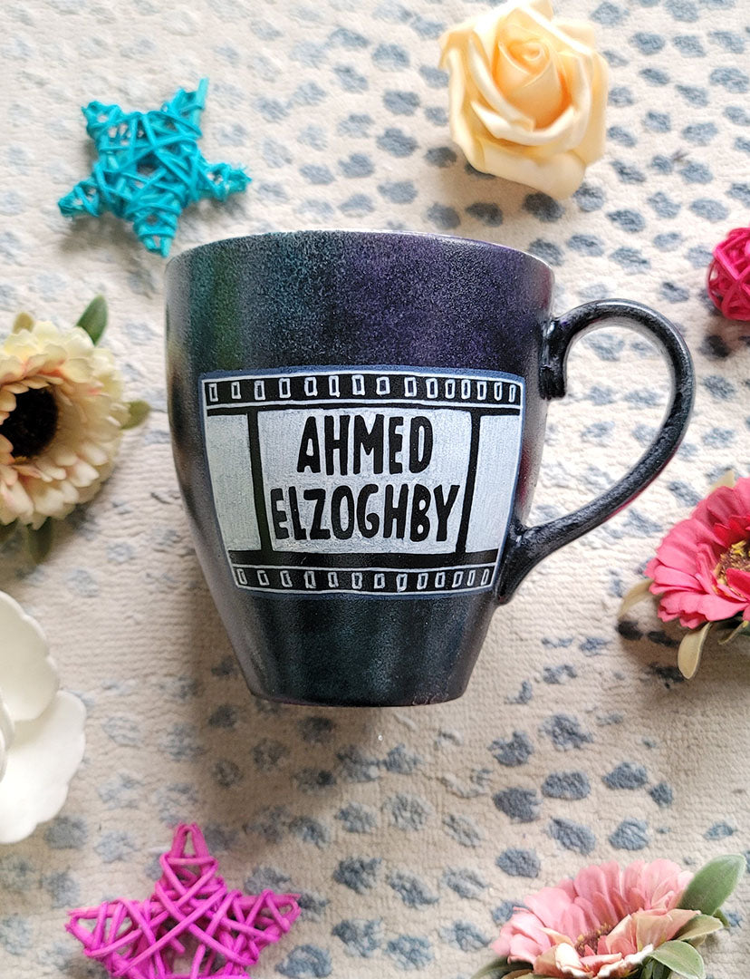 A dark sky mug with part of a film roll with the name "Ahmed El Zoghby" written on it