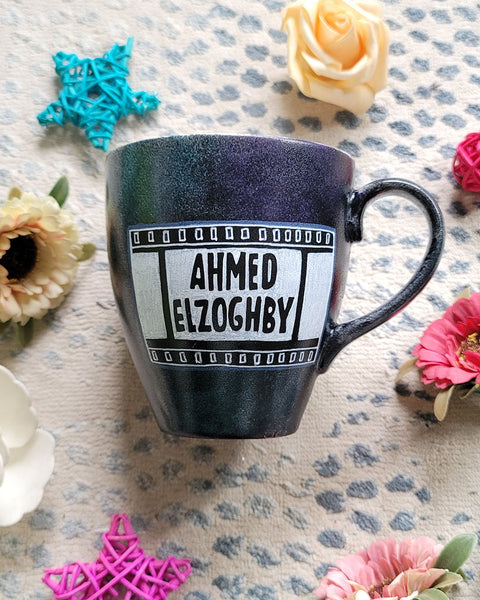 A dark sky mug with part of a film roll with the name "Ahmed El Zoghby" written on it