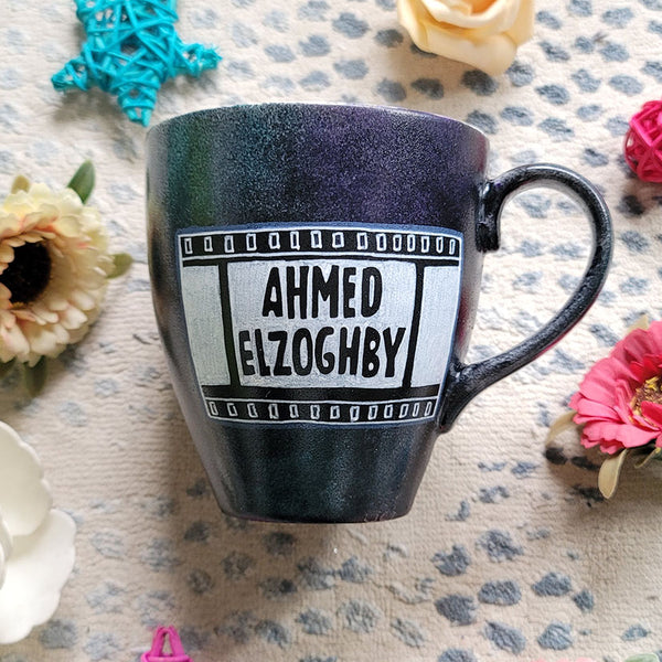 A dark sky mug with part of a film roll with the name "Ahmed El Zoghby" written on it