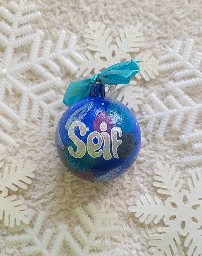 A Colorful hand painted ornament with blue background and colors of blue, black dark purple and silver scattered all over with white dots and the name "seif" written in silver outlined with white