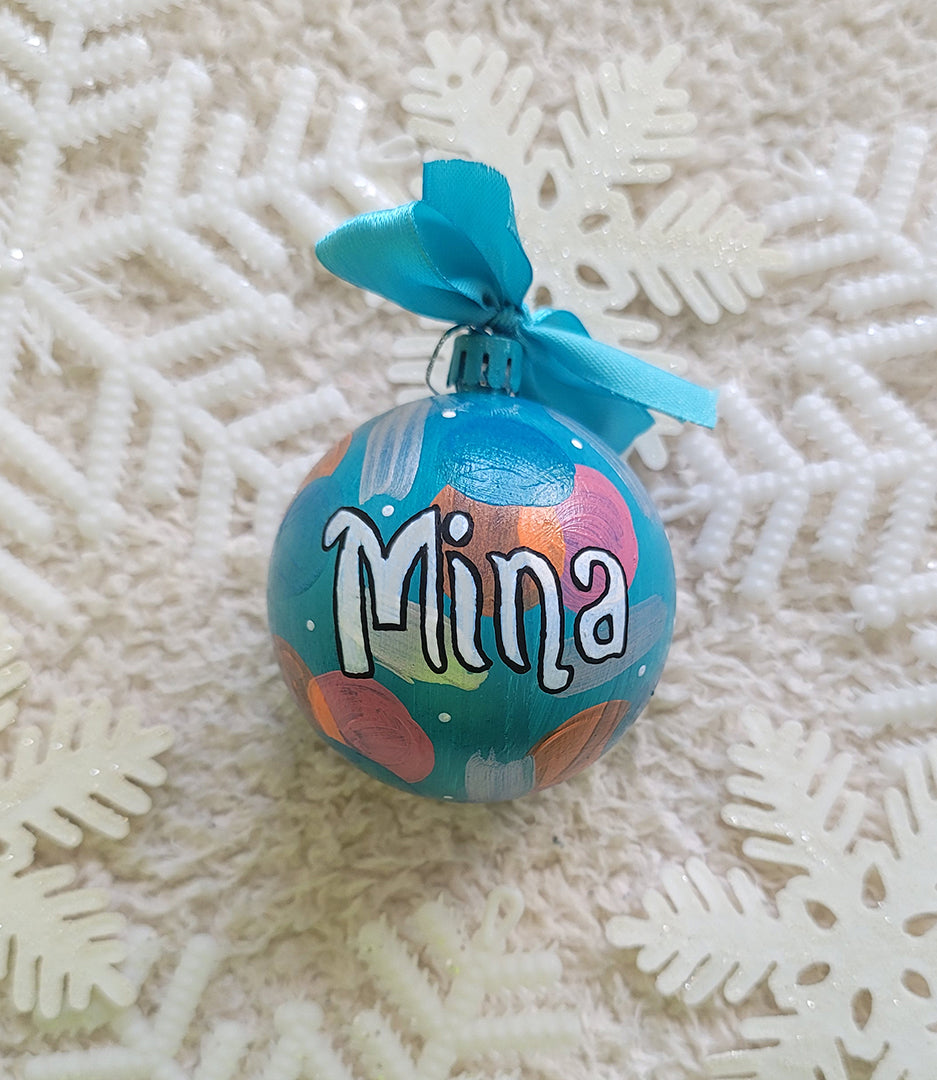 A Colorful hand painted ornament with turquoise bubbly background and colors of bubbly, pastel orange, pastel pink and silver scattered all over with white dots and the name "Mina" written in white outlined with black