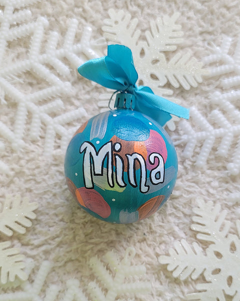 A Colorful hand painted ornament with turquoise bubbly background and colors of bubbly, pastel orange, pastel pink and silver scattered all over with white dots and the name "Mina" written in white outlined with black