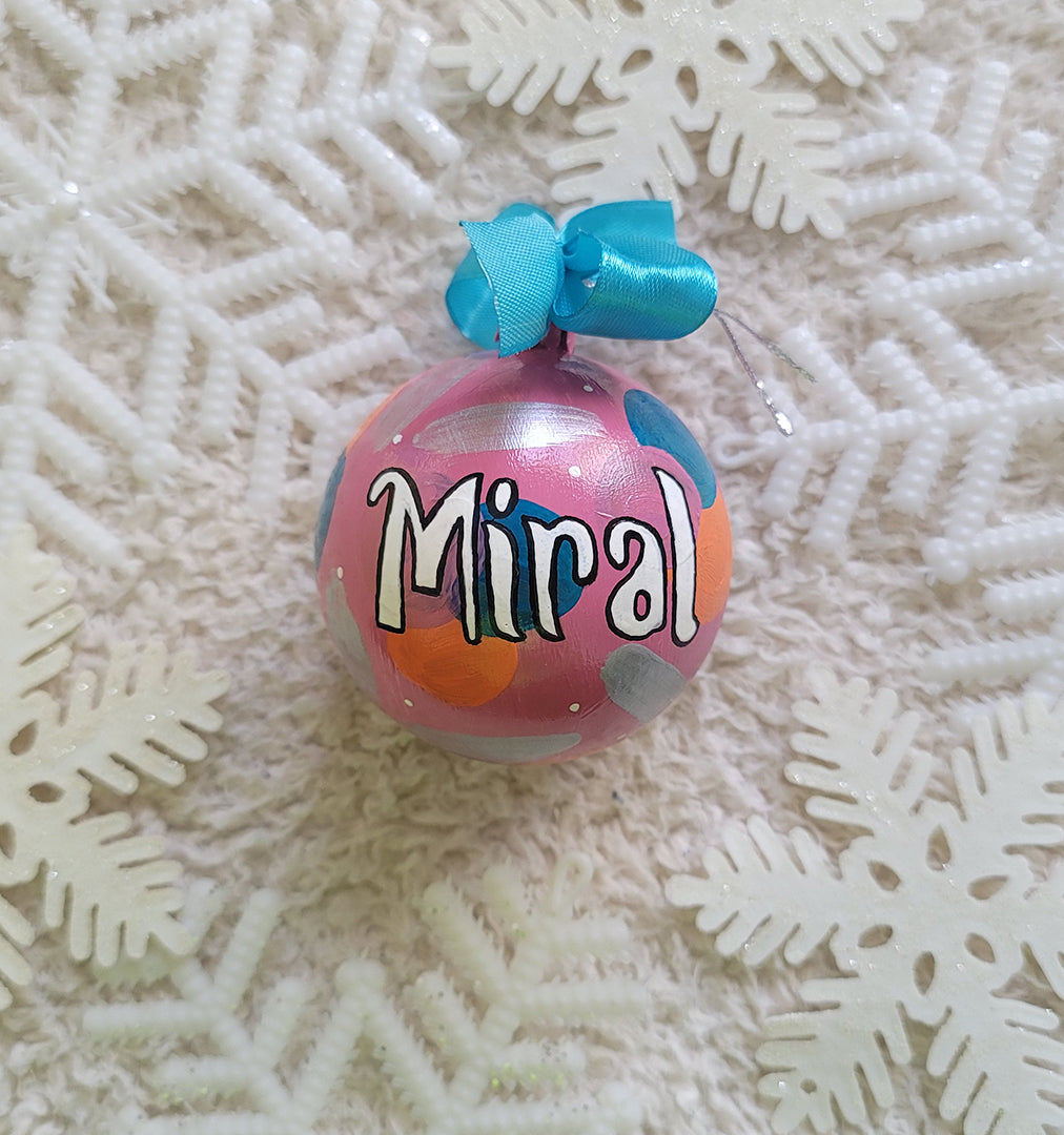 A Colorful hand painted ornament with pastel pink background and colors of pastel pink, teal orange and silver scattered all over with white dots and the name "Miral" written in white outlined with black