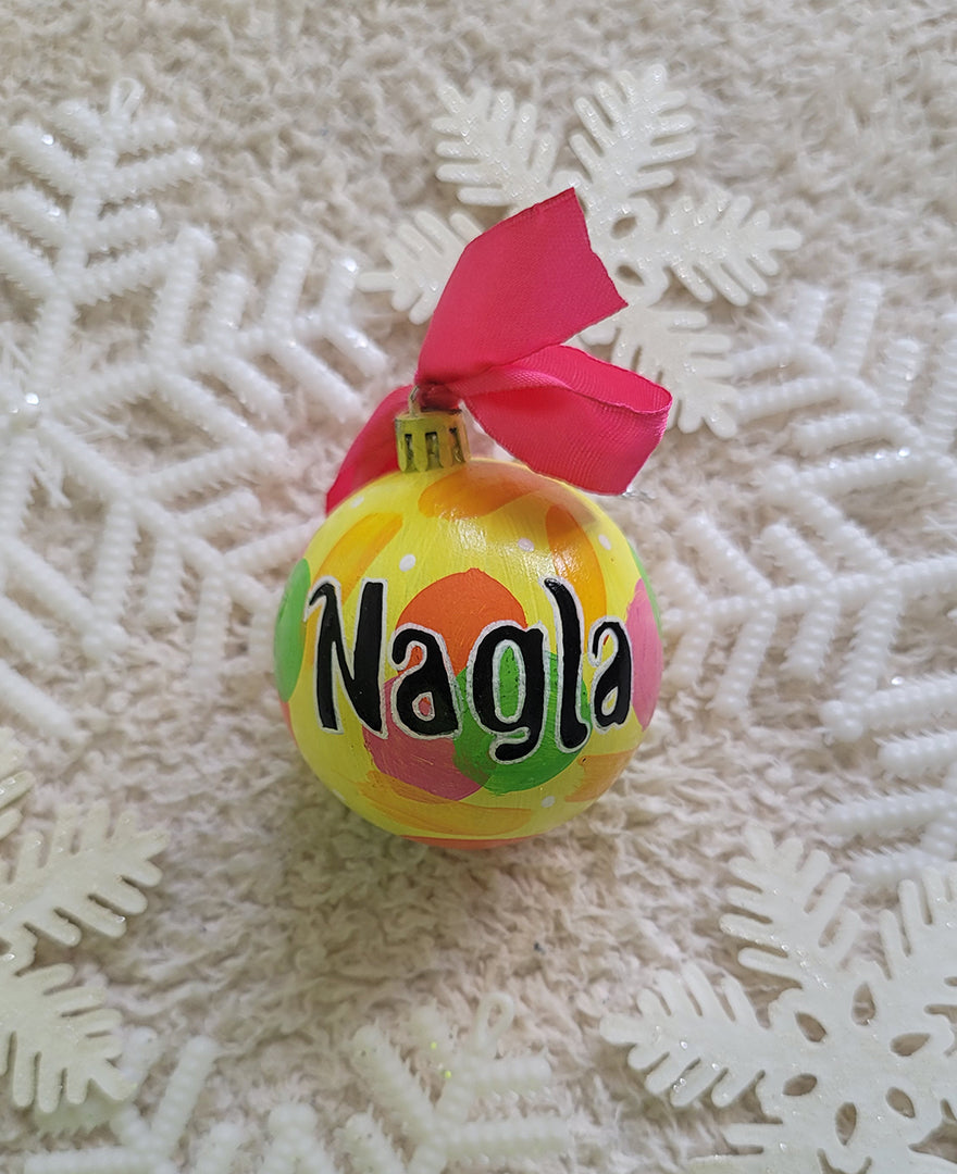 A Colorful hand painted ornament with yellow background and colors of pink, orange, green and yellow scattered all over with white dots and the name "Nagla" written in black outlined with white