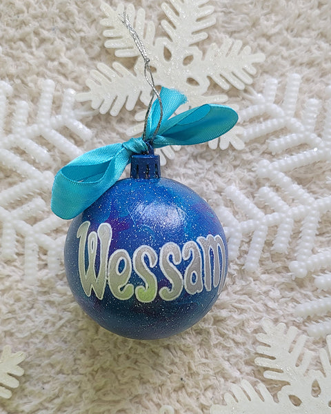 A Blue galaxy hand painted ornament with the name "Wessam" written in silver outlined with white