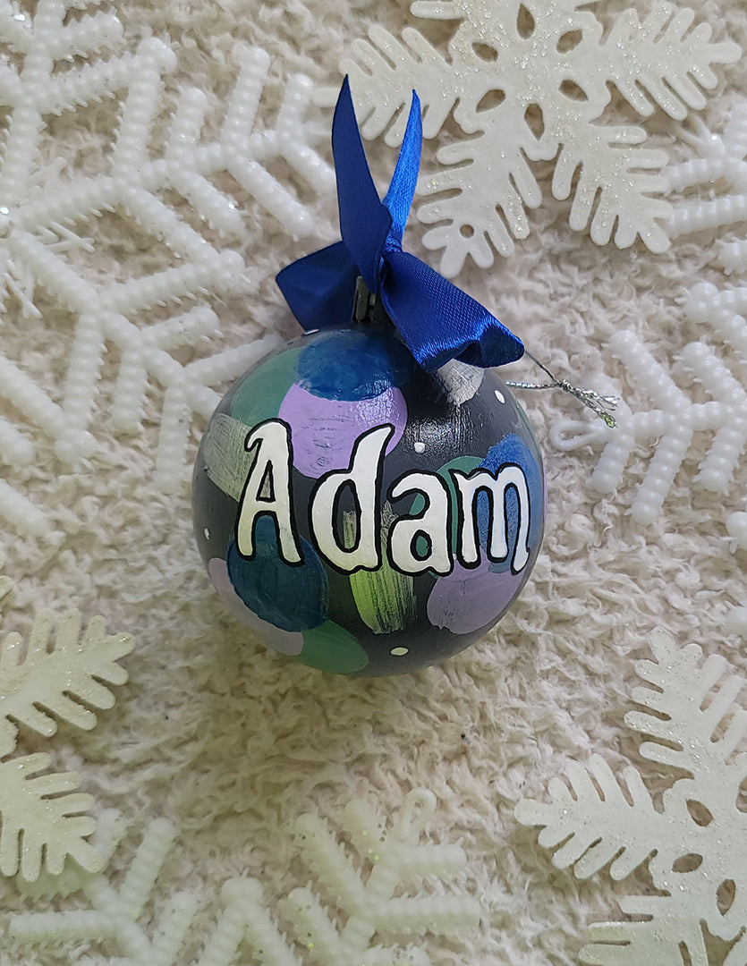 A Colorful hand painted ornament with Grey background and colors of lilac, blue, green and silver scattered all over with white dots and the name "Adam" written in white outlined with black