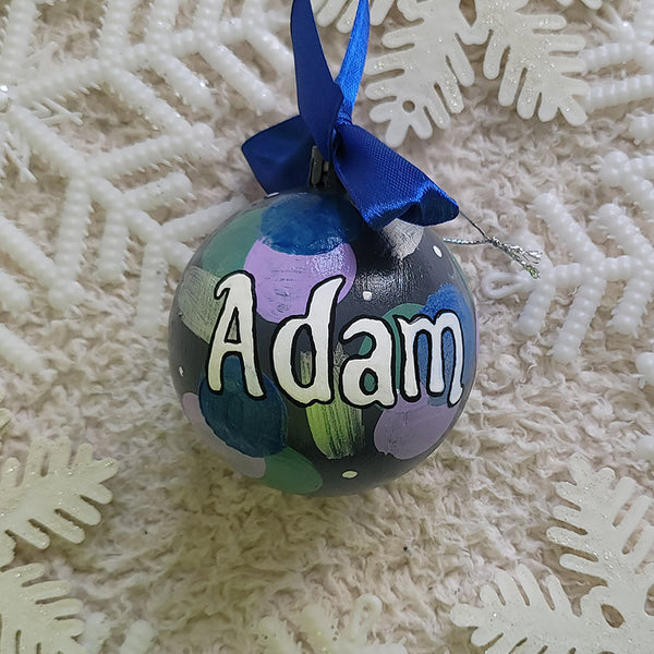 A Colorful hand painted ornament with Grey background and colors of lilac, blue, green and silver scattered all over with white dots and the name "Adam" written in white outlined with black