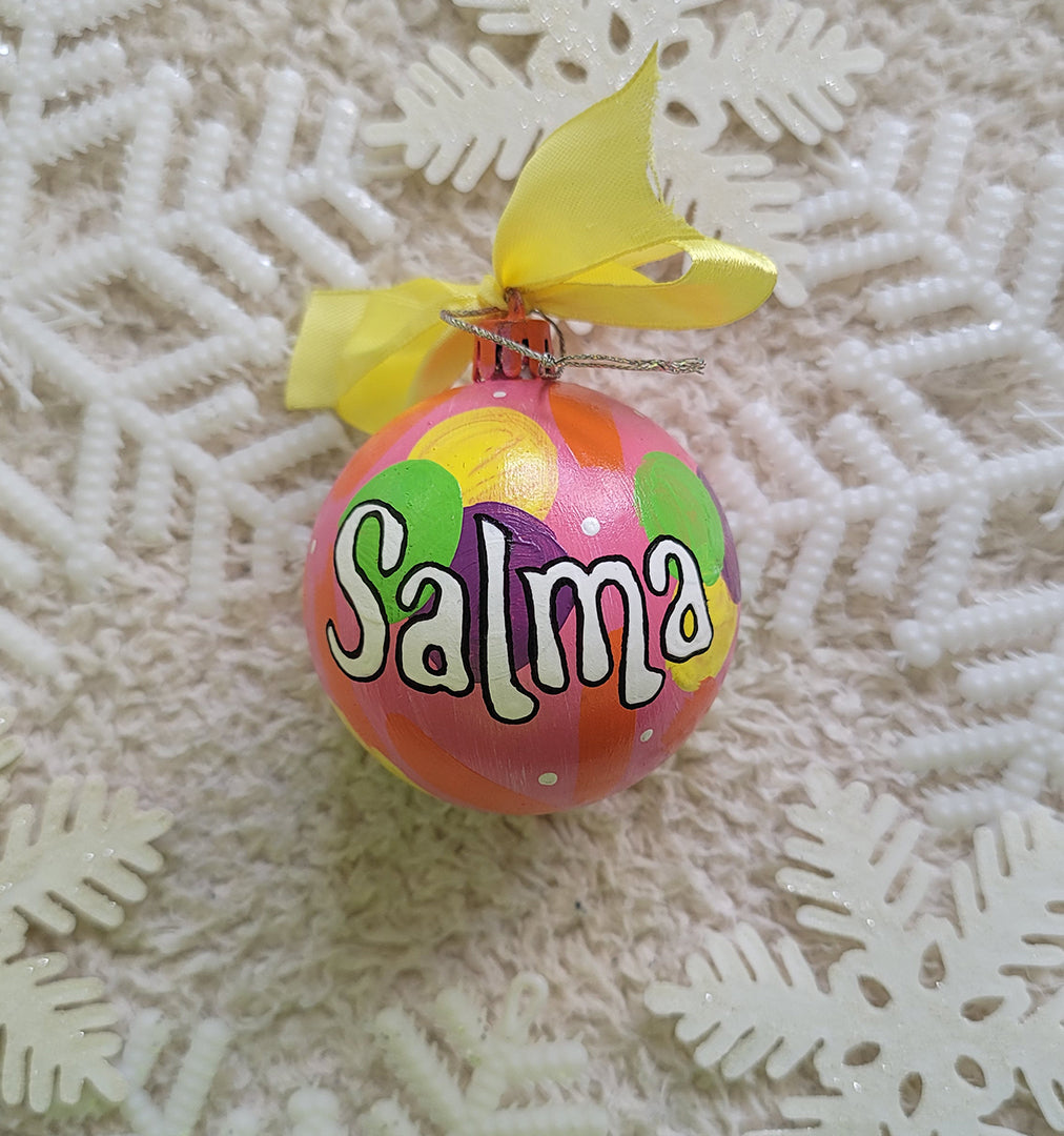 A Colorful hand painted ornament with Pink background and colors of pink, purple, green and yellow scattered all over with white dots and the name "Salma" written in white outlined with black