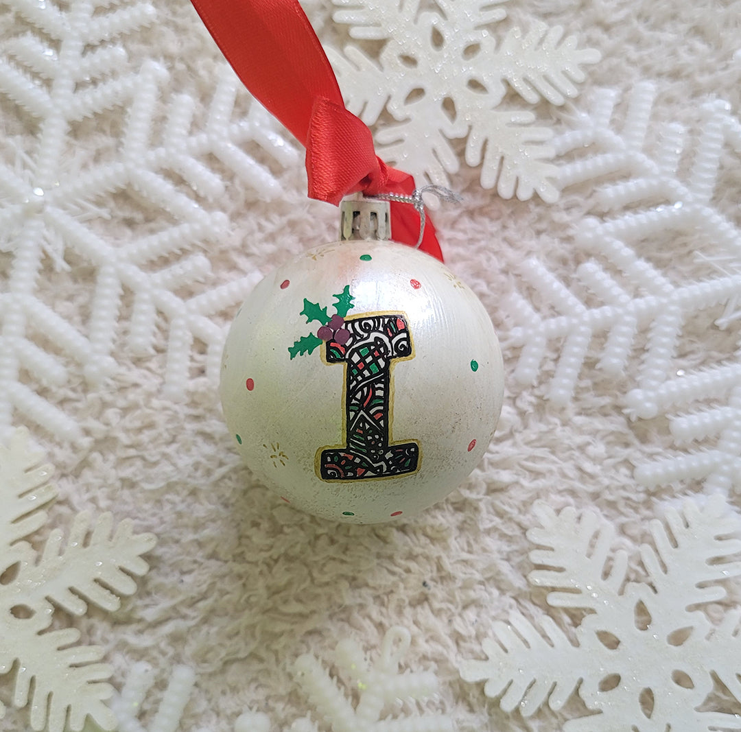 A Pearl white hand painted ball ornament with the letter "I" drawn filled with black doodles and colored with green and red, outlined with gold and green  and red dots scattered all over the ball with gold stars