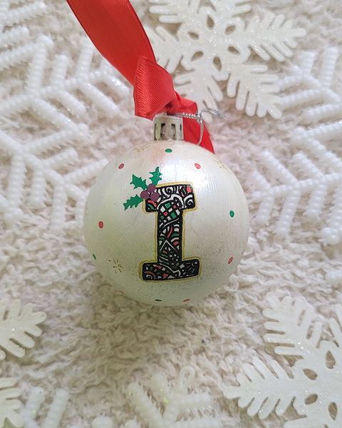 A Pearl white hand painted ball ornament with the letter "I" drawn filled with black doodles and colored with green and red, outlined with gold and green  and red dots scattered all over the ball with gold stars