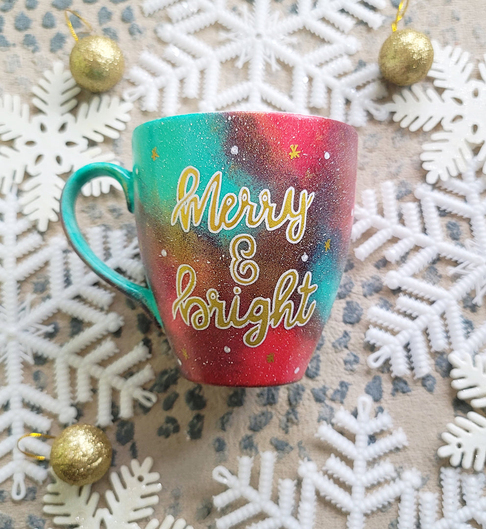 A Christmas galaxy colored hand painted mug with the words "Merry and bright" written gold with white dots and gold stars scattered all over