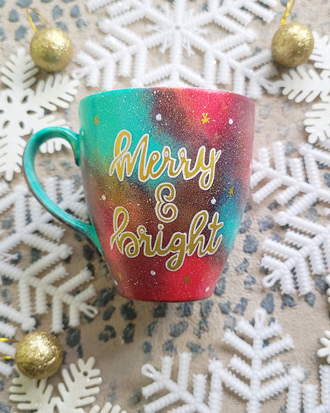 A Christmas galaxy colored hand painted mug with the words "Merry and bright" written gold with white dots and gold stars scattered all over