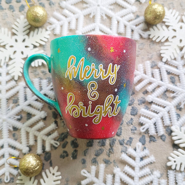 A Christmas galaxy colored hand painted mug with the words "Merry and bright" written gold with white dots and gold stars scattered all over