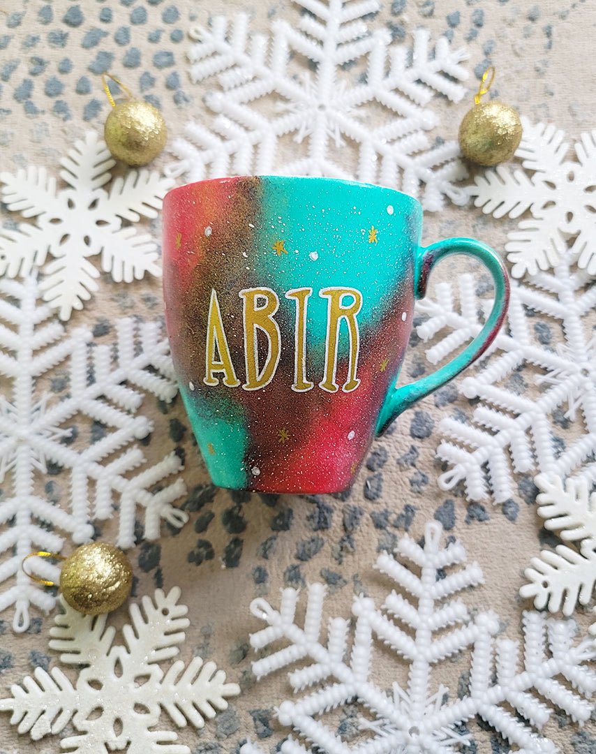 A Christmas galaxy colored hand painted mug with the name "Abir" written gold with white dots and gold stars scattered all over