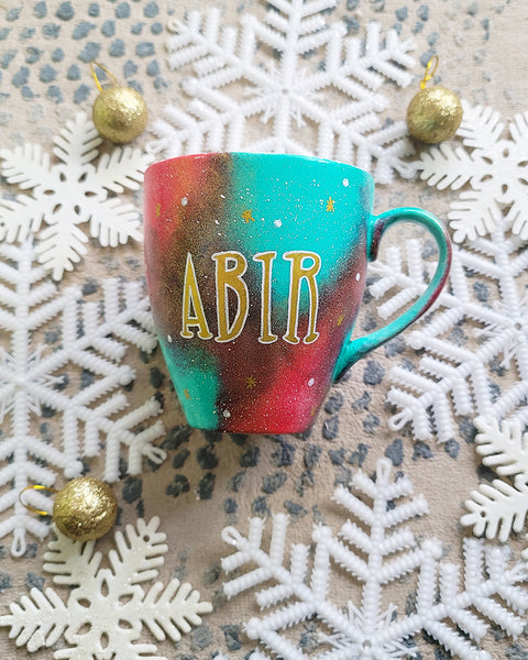 A Christmas galaxy colored hand painted mug with the name "Abir" written gold with white dots and gold stars scattered all over