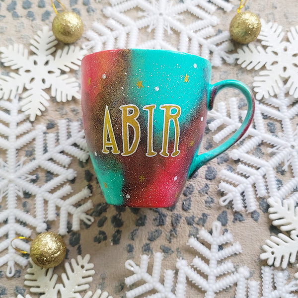 A Christmas galaxy colored hand painted mug with the name "Abir" written gold with white dots and gold stars scattered all over