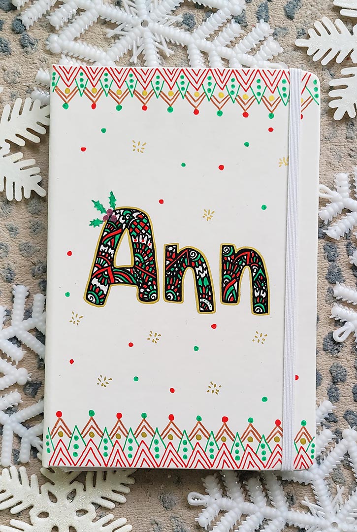 A Pearl white notebook with the letters "ANN" written filled with doodles in black and colored in green and red. then hz patterns drawn all through the bottom of the mug. with green and red dots, and gold stars scattered all over