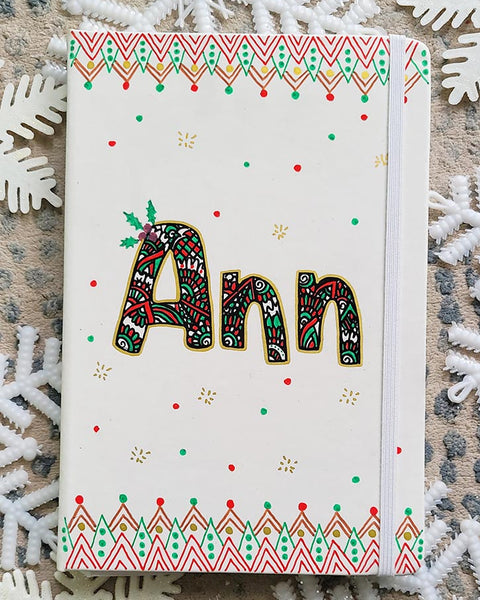 A Pearl white notebook with the letters "ANN" written filled with doodles in black and colored in green and red. then hz patterns drawn all through the bottom of the mug. with green and red dots, and gold stars scattered all over