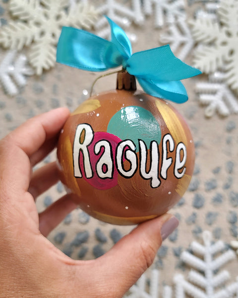 A Colorful hand painted ornament with choco brown background and colors of choco brown, indian pink, mint green and gold scattered all over with white dots and the name "Raoufe" written in white outlined with black