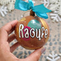 A Colorful hand painted ornament with choco brown background and colors of choco brown, indian pink, mint green and gold scattered all over with white dots and the name "Raoufe" written in white outlined with black