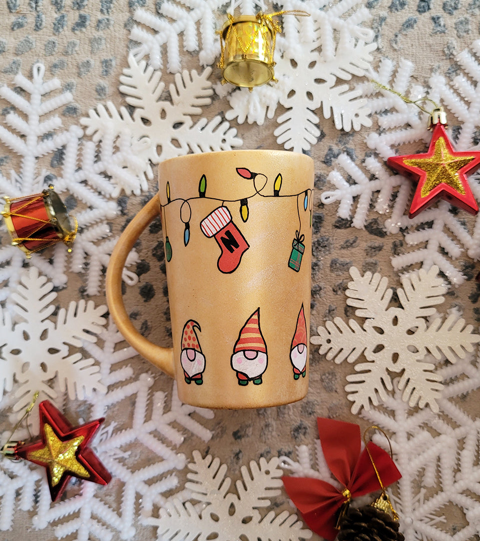 A Gold hand painted mug with gnomes all around the bottom half of the mug and festive lighting all over the top of the mug and a stocking hung from the lights with the letter N