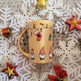 A Gold hand painted mug with gnomes all around the bottom half of the mug and festive lighting all over the top of the mug and a stocking hung from the lights with the letter N