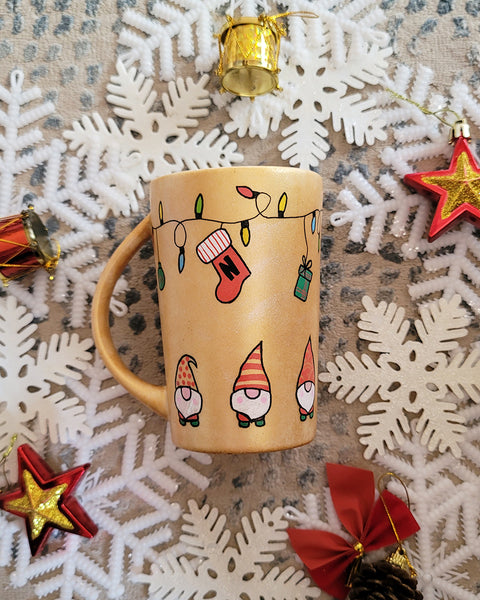 A Gold hand painted mug with gnomes all around the bottom half of the mug and festive lighting all over the top of the mug and a stocking hung from the lights with the letter N