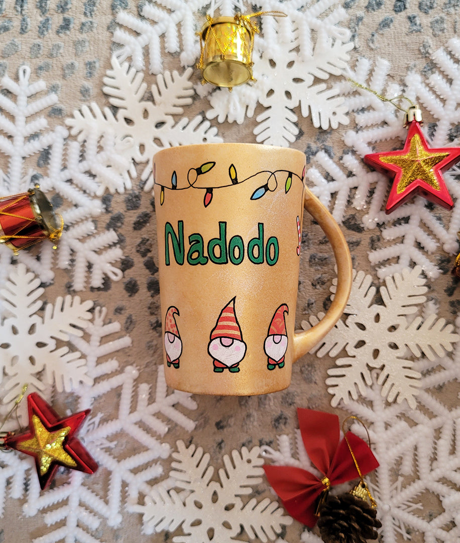 A Gold hand painted mug with gnomes all around the bottom half of the mug and festive lighting all over the top of the mug and the name nadodo in the center