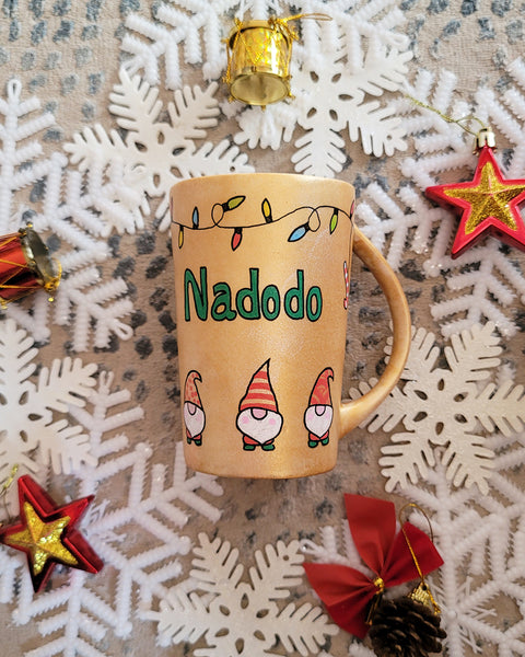 A Gold hand painted mug with gnomes all around the bottom half of the mug and festive lighting all over the top of the mug and the name nadodo in the center