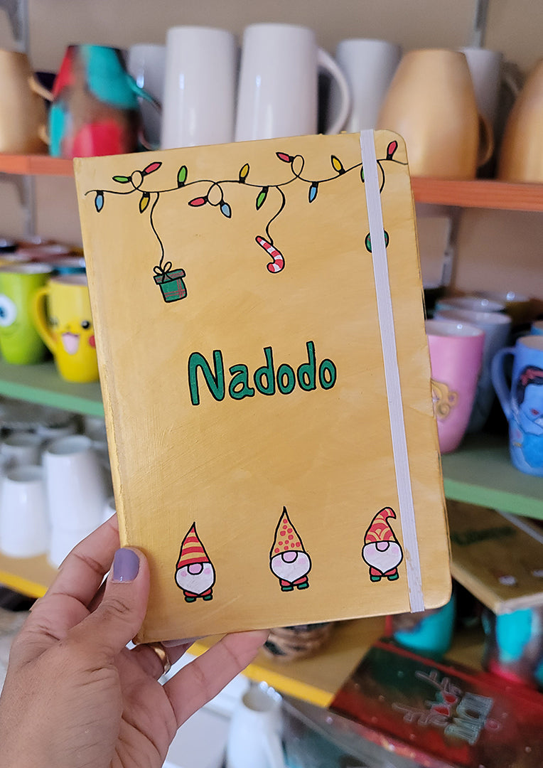 A Gold hand painted A5 Notebook with gnomes on the bottom and festive light on the top and the name "Nadodo" written in the middle in green