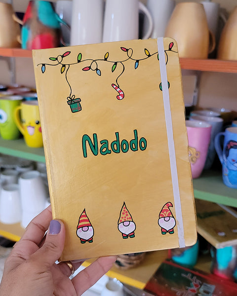 A Gold hand painted A5 Notebook with gnomes on the bottom and festive light on the top and the name "Nadodo" written in the middle in green