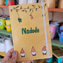 A Gold hand painted A5 Notebook with gnomes on the bottom and festive light on the top and the name "Nadodo" written in the middle in green