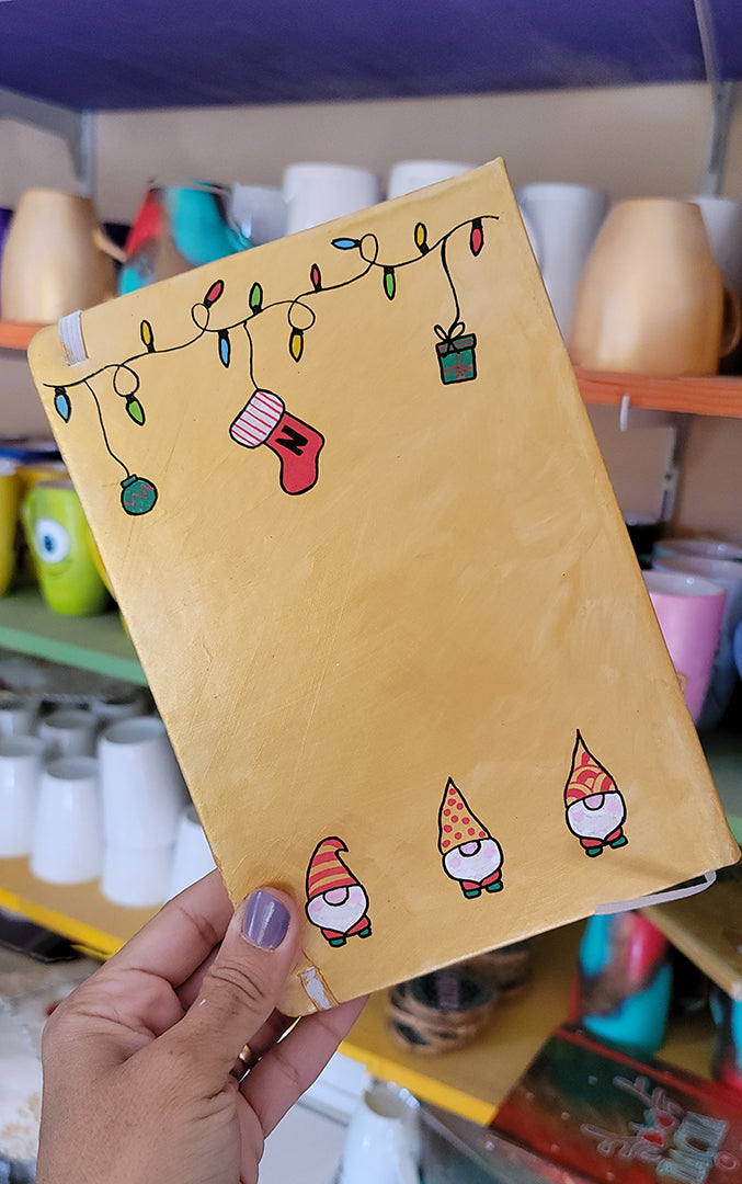 A Gold hand painted A5 Notebook with gnomes on the bottom and festive light on the top and a red stocking  with the letter N hanging from the festive lights