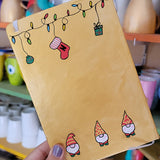 A Gold hand painted A5 Notebook with gnomes on the bottom and festive light on the top and a red stocking  with the letter N hanging from the festive lights