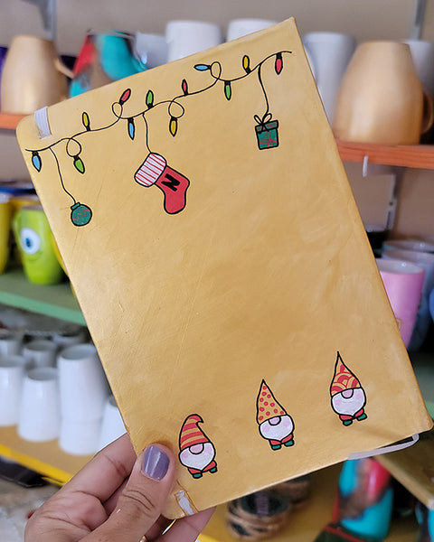 A Gold hand painted A5 Notebook with gnomes on the bottom and festive light on the top and a red stocking  with the letter N hanging from the festive lights