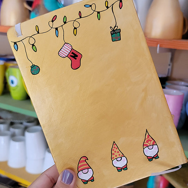 A Gold hand painted A5 Notebook with gnomes on the bottom and festive light on the top and a red stocking  with the letter N hanging from the festive lights