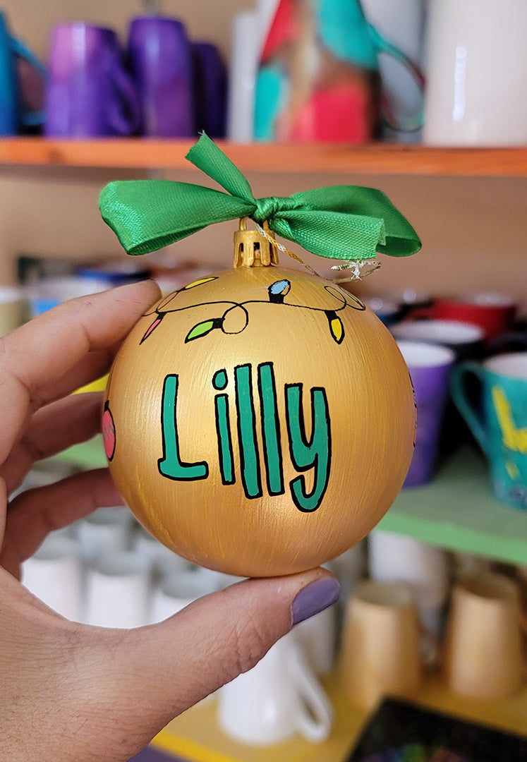 A gold hand painted ball ornament with festive lights all around the top and the name "Lilly" written in green outlined with black