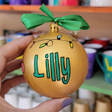 A gold hand painted ball ornament with festive lights all around the top and the name 