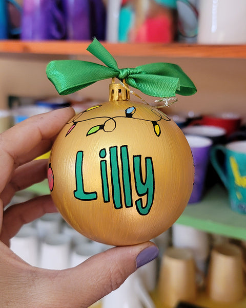 A gold hand painted ball ornament with festive lights all around the top and the name "Lilly" written in green outlined with black