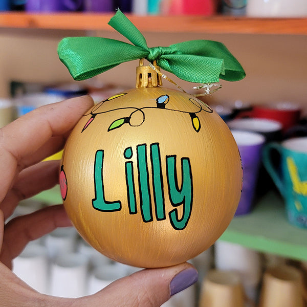 A gold hand painted ball ornament with festive lights all around the top and the name "Lilly" written in green outlined with black