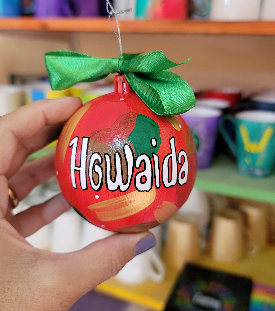 A Colorful hand painted ornament with red background and colors of red green, brown and gold scattered all over with white dots and the name "Howaida" written in white outlined with black