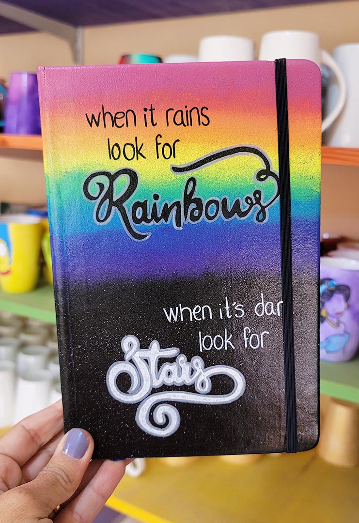 A notebook with the top half painted in rainbow and the bottom in dark galaxy, with the words on the rainbow written "when it rains look for rainbows" and the words on the galaxy written "when its dark look for stars"