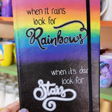 A notebook with the top half painted in rainbow and the bottom in dark galaxy, with the words on the rainbow written 