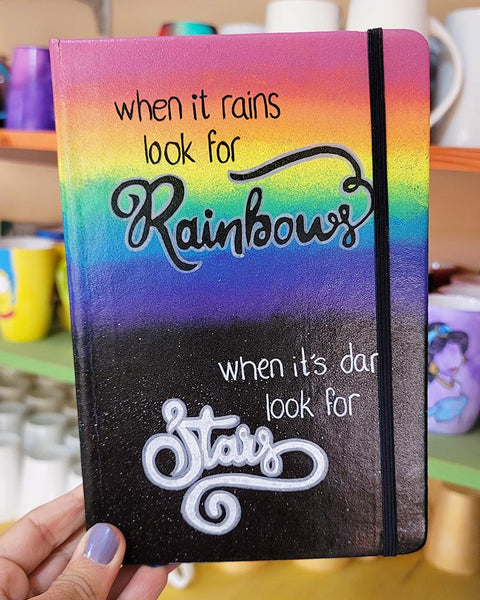 A notebook with the top half painted in rainbow and the bottom in dark galaxy, with the words on the rainbow written "when it rains look for rainbows" and the words on the galaxy written "when its dark look for stars"