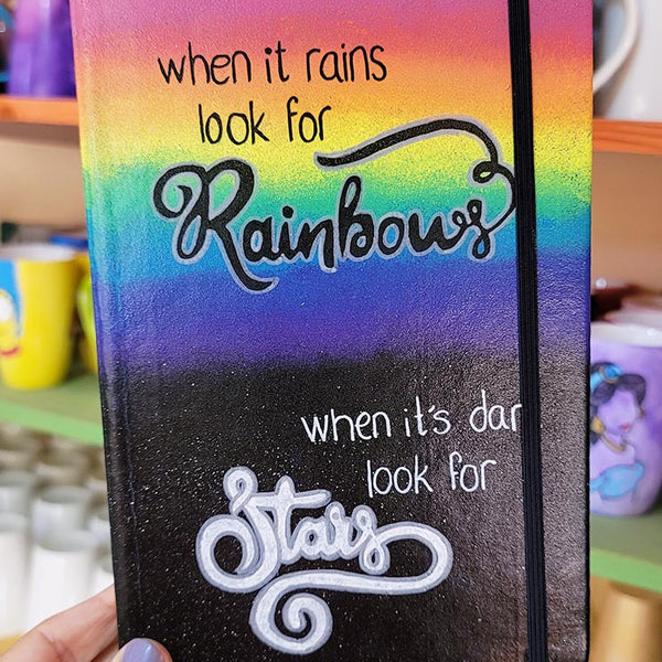 A notebook with the top half painted in rainbow and the bottom in dark galaxy, with the words on the rainbow written "when it rains look for rainbows" and the words on the galaxy written "when its dark look for stars"