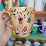 A Gold hand painted mug with gnomes all around the bottom half of the mug and festive lighting all over the top of the mug and a stocking hung from the lights with the letter M