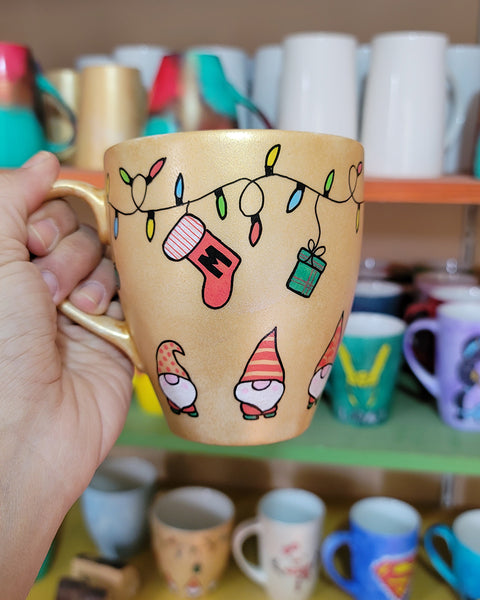 A Gold hand painted mug with gnomes all around the bottom half of the mug and festive lighting all over the top of the mug and a stocking hung from the lights with the letter M