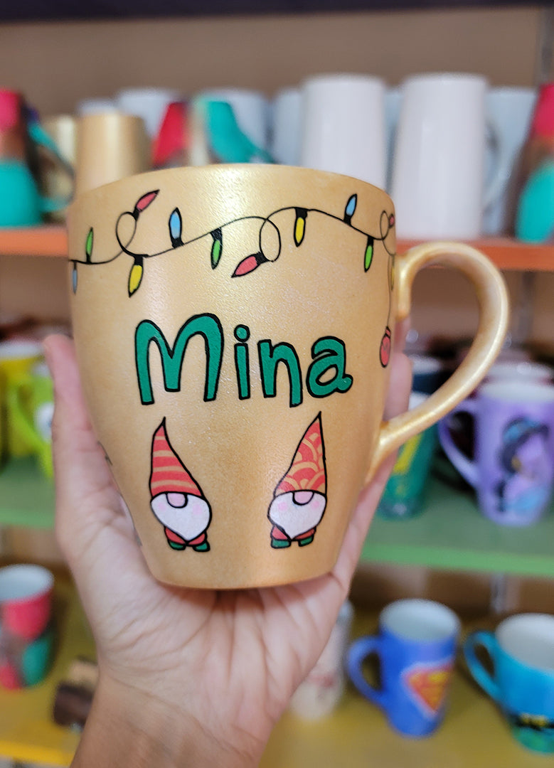 A Gold hand painted mug with gnomes all around the bottom half of the mug and festive lighting all over the top of the mug and the name Mina in the center