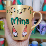 A Gold hand painted mug with gnomes all around the bottom half of the mug and festive lighting all over the top of the mug and the name Mina in the center